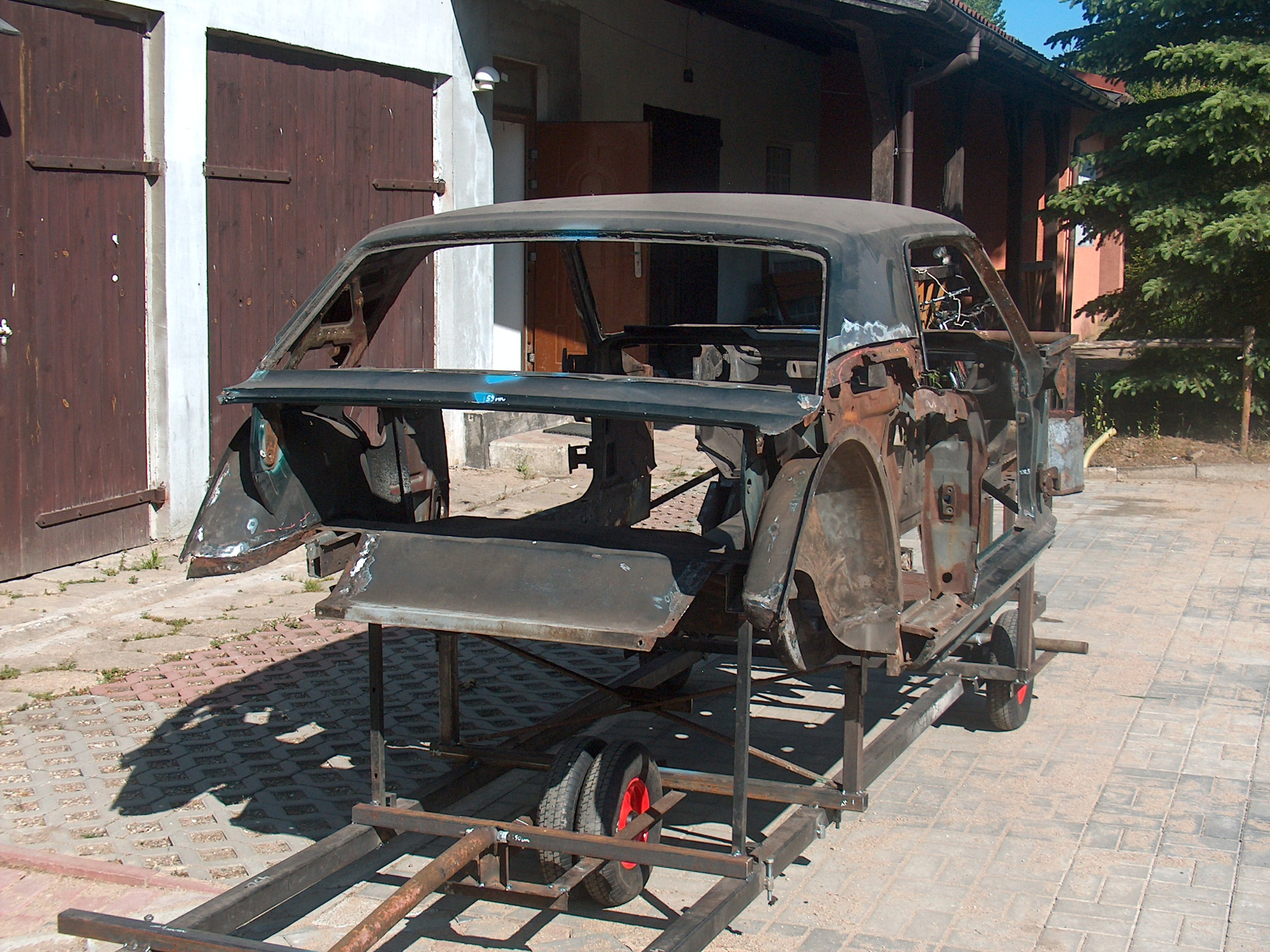 Goły szkielet karoserii klasycznego auta na metalowym stelażu z kołami, przygotowany do piaskowania lub malowania proszkowego na zewnątrz budynku.