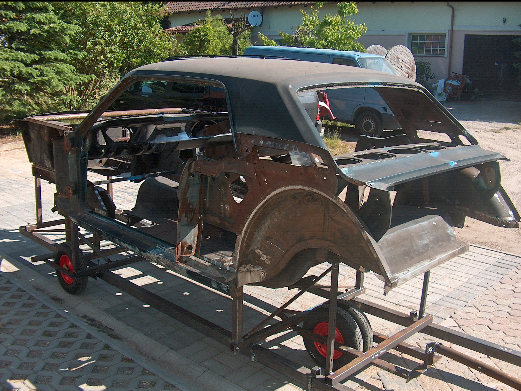 Karoseria klasycznego auta na metalowym stelażu z czerwonymi kołami, w trakcie renowacji. Widoczne ślady rdzy i przygotowania do dalszych prac.