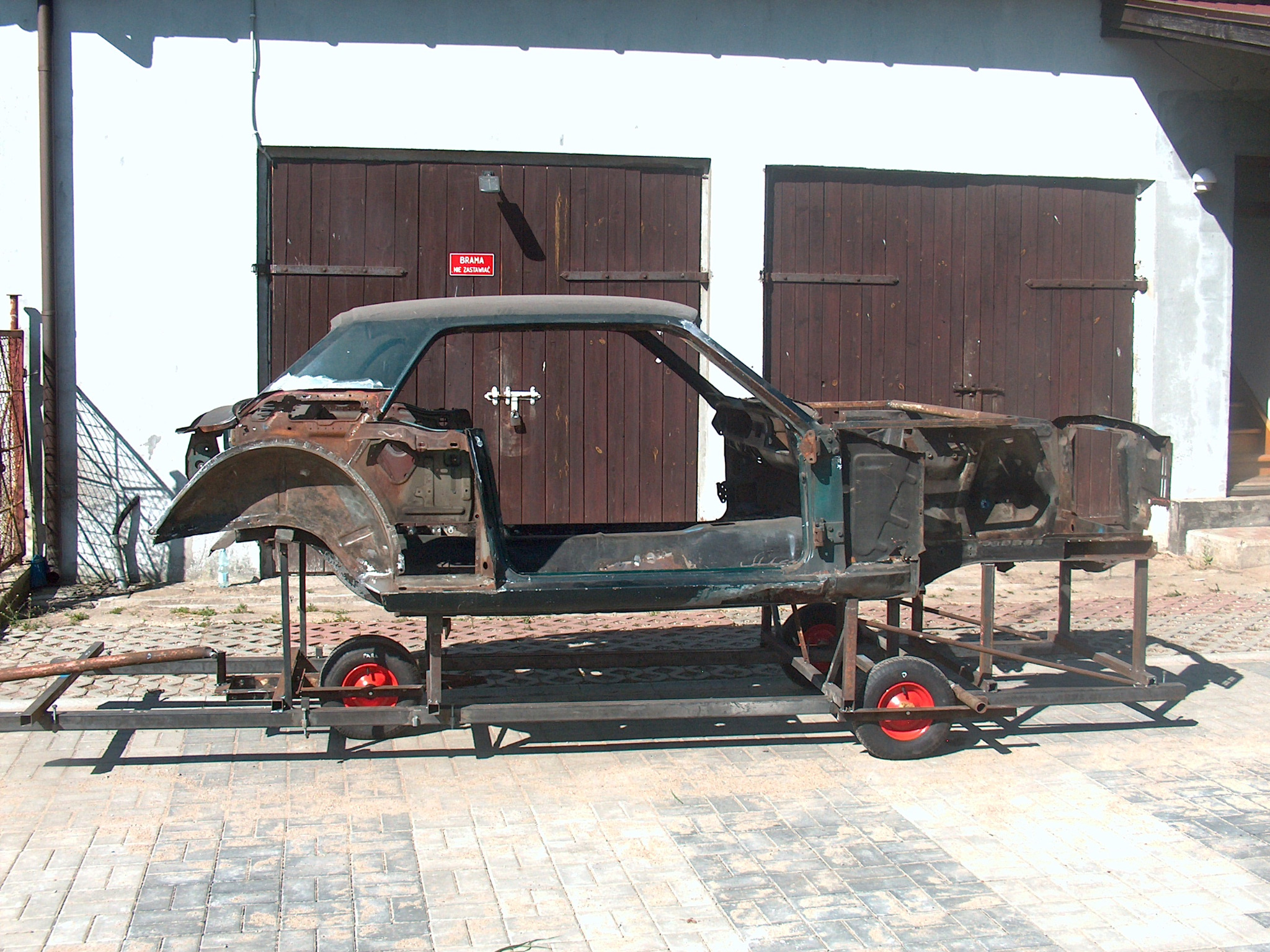 Karoseria klasycznego auta na wózku warsztatowym, przygotowana do renowacji. Widoczne ślady rdzy i demontażu elementów. Tło stanowi budynek z drewnianymi bramami.