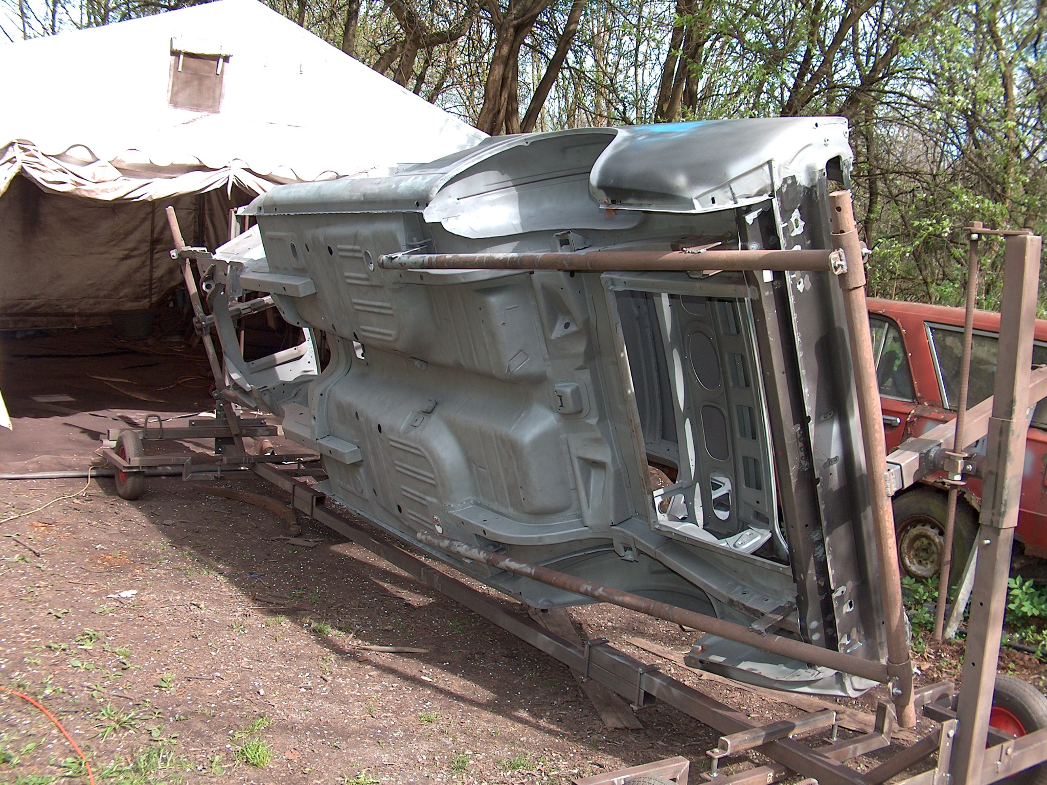 Karoseria samochodu po renowacji, zamontowana na specjalnym stelażu, w tle widoczny namiot i drzewa. Elementy konstrukcyjne pojazdu w szarym kolorze.
