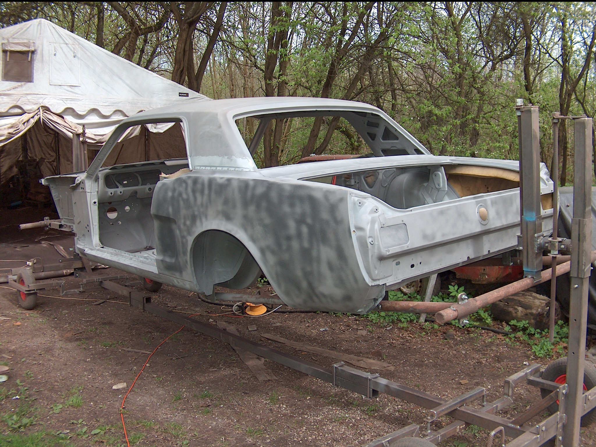 Nadwozie klasycznego Forda Mustanga, osadzone na metalowej konstrukcji w trakcie prac renowacyjnych. Widoczne ślady szpachli i podkładu, tło z zielenią i namiotem.