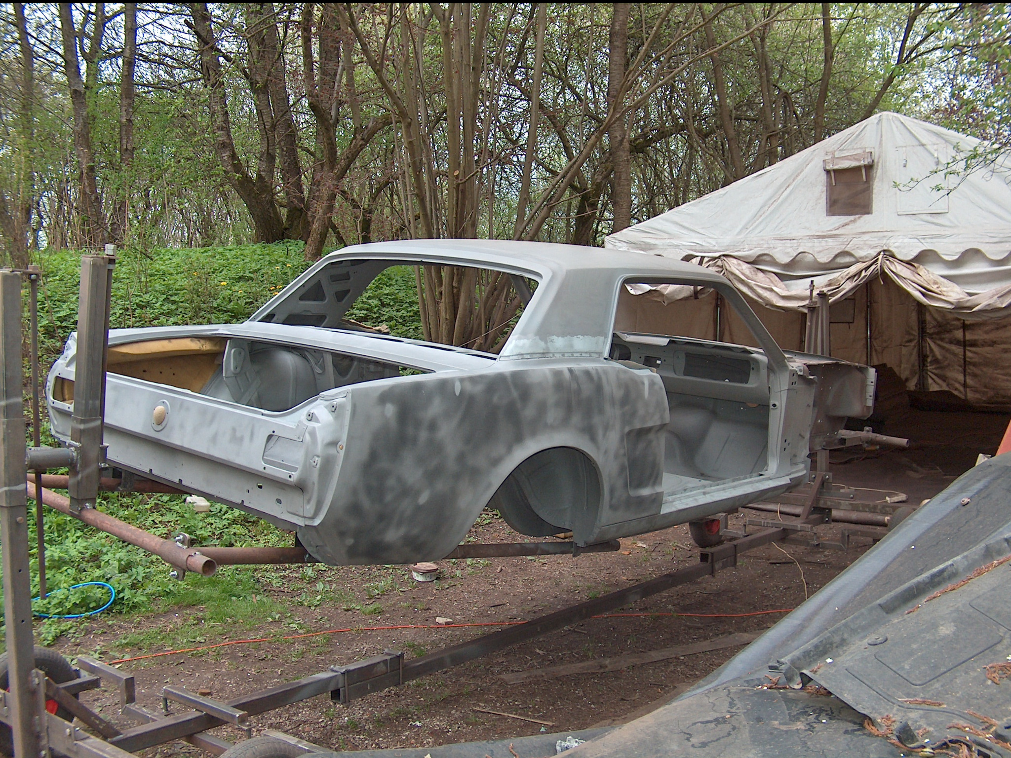 Karoseria klasycznego auta, częściowo oczyszczona, zawieszona na metalowej konstrukcji w plenerze. W tle widoczny namiot i zieleń.