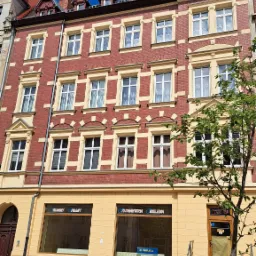 Odnowiona kamienica w Zgorzelcu z czerwoną cegłą, kremowymi zdobieniami okien i niebieskim dachem wieżyczki, widok z poziomu ulicy w słoneczny dzień.