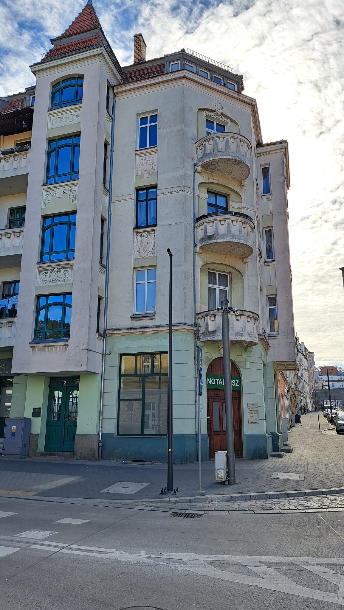 Kamienica narożna z balkonami, zielonymi oknami i szyldem 'Notariusz' w słoneczny dzień.