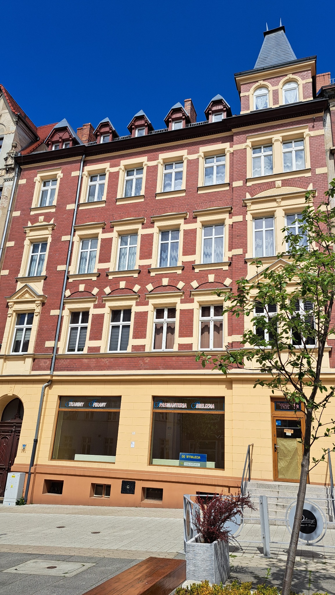 Odnowiona kamienica w Zgorzelcu z czerwoną cegłą, kremowymi zdobieniami okien i niebieskim dachem wieżyczki, widok z poziomu ulicy w słoneczny dzień.