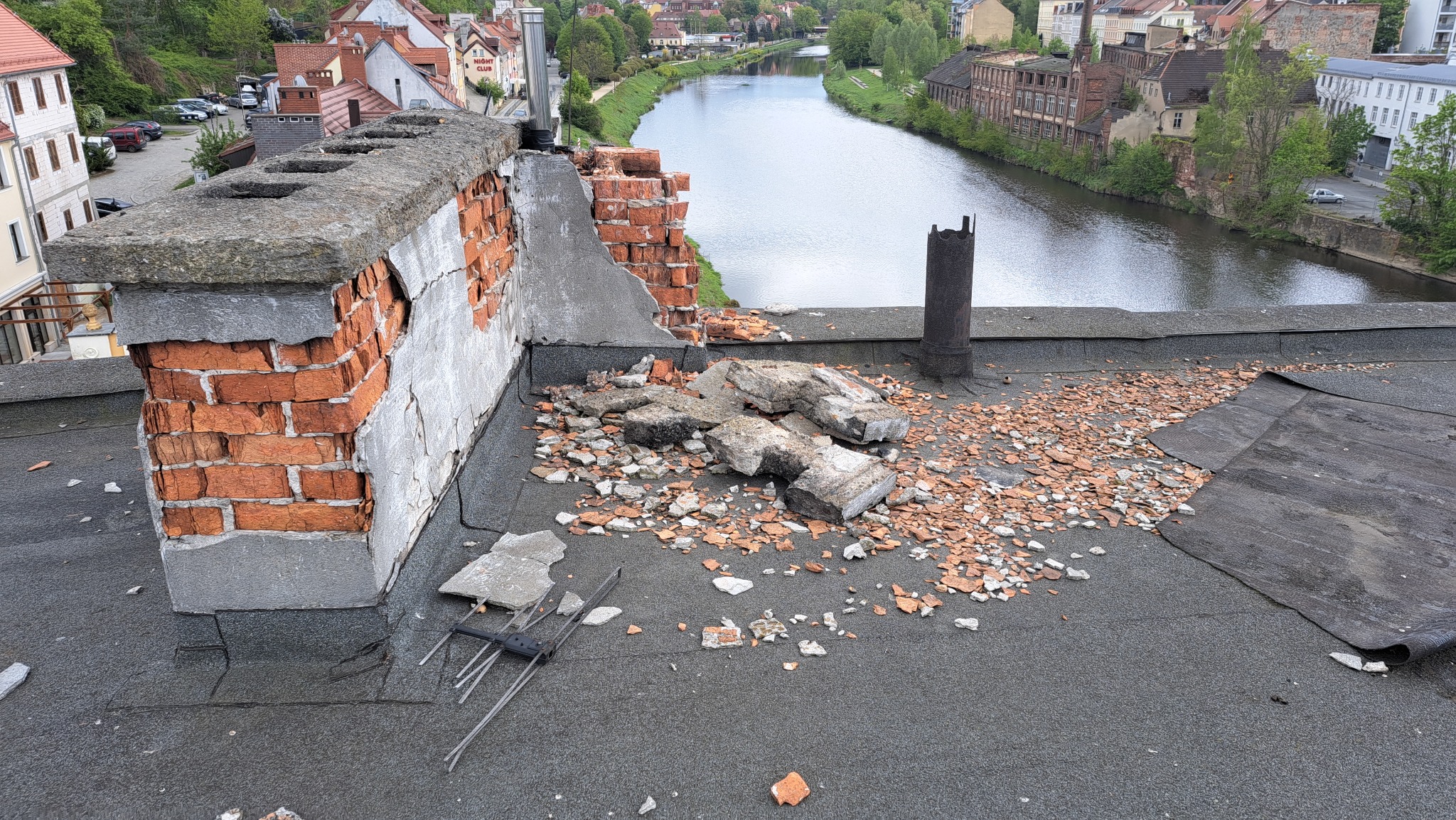 Uszkodzony komin z cegły na dachu budynku z widokiem na rzekę i zabudowę miejską, gruz i fragmenty cegieł rozsypane wokół, widoczna antena telewizyjna.