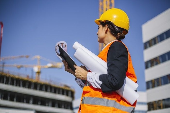 Kobieta w żółtym kasku i pomarańczowej kamizelce, trzymająca zwinięte plany i czarną podkładkę, stojąca na tle budowy z dźwigiem i nowoczesnym budynkiem.