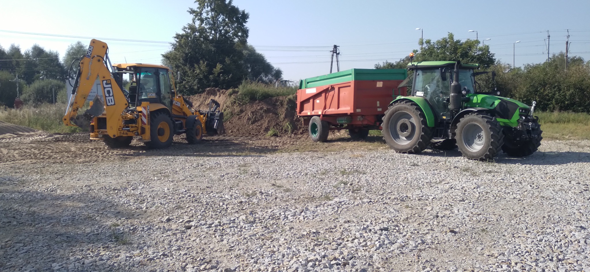 Żółta koparko-ładowarka JCB i zielony ciągnik z czerwoną przyczepą na żwirowym terenie, w tle niewielki nasyp ziemi i zieleń.