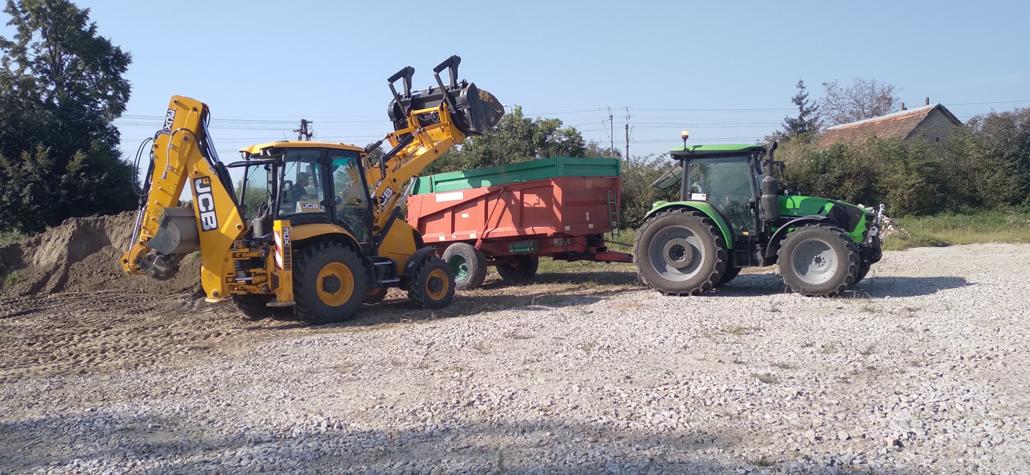 Żółta koparko-ładowarka JCB na żwirowym podłożu, załadowująca ziemię do czerwono-zielonej przyczepy ciągniętej przez zielony traktor Deutz-Fahr, w tle drzewa i budynek.