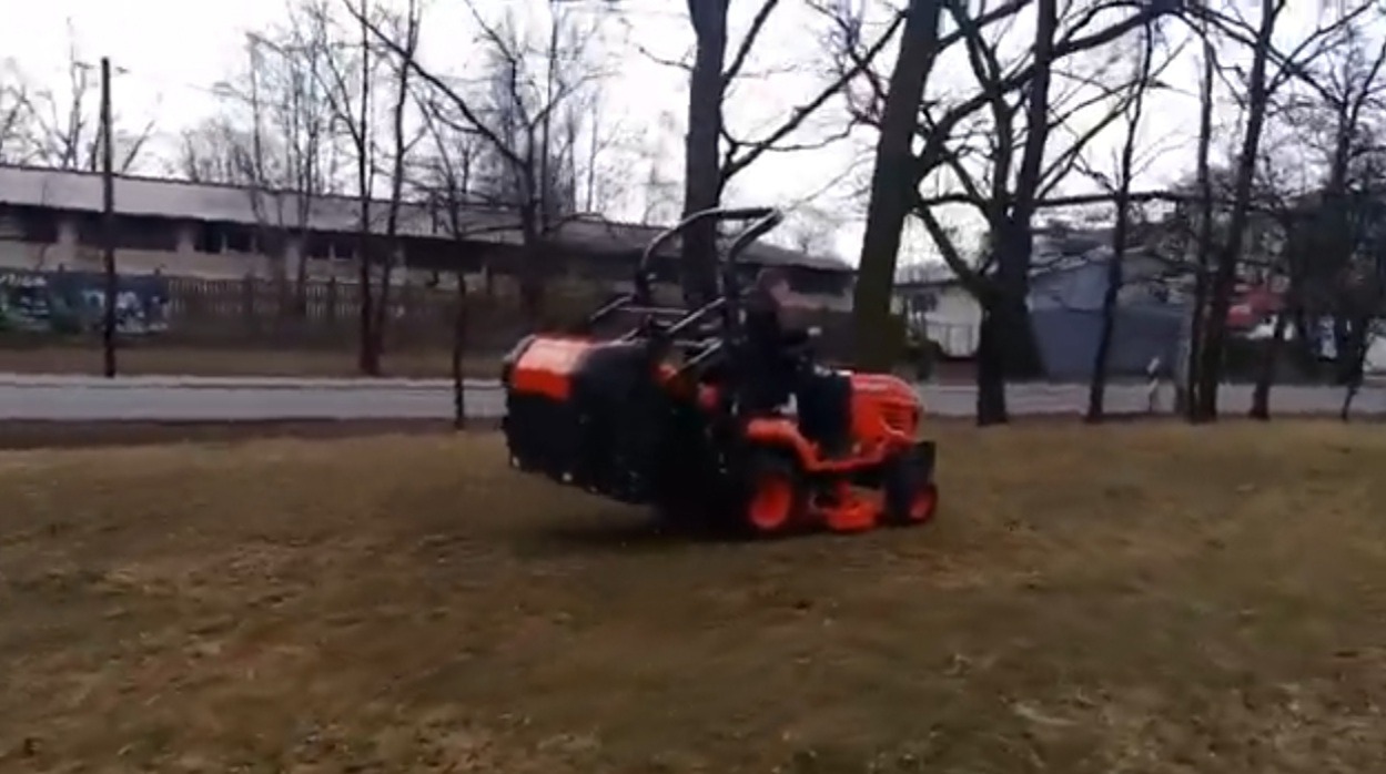 Pomarańczowy traktor z czarnym kosiarką rotacyjną na nierównym trawniku, w tle budynek i drzewa bez liści.