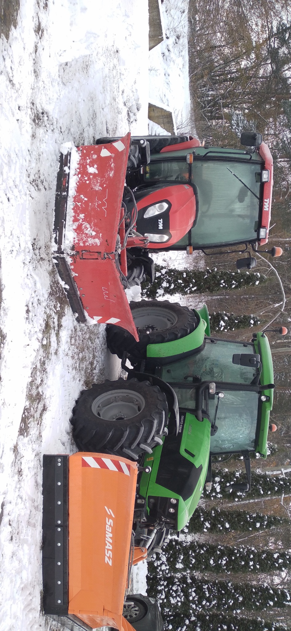 Dwa traktory z pługami do śniegu, czerwony i zielony, stojące na ośnieżonym terenie, z widocznym lasem w tle.