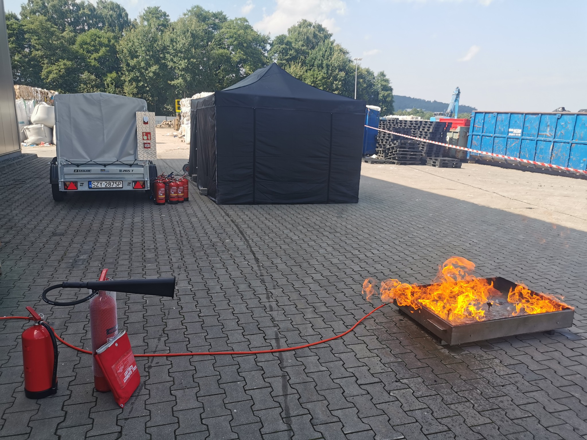 Symulacja pożaru w metalowej wannie podczas szkolenia w Lipowej, widoczne gaśnice, przyczepka oraz czarny namiot w tle.