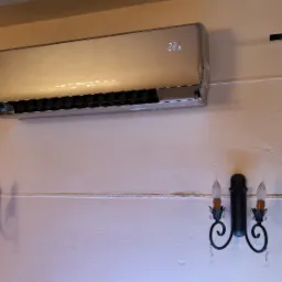 Srebrny klimatyzator z wyświetlaczem temperatury 22,5 stopni Celsjusza zamontowany na kremowej ścianie, oświetlony przez dwa kinkiety w stylu retro z imitacjami świec.