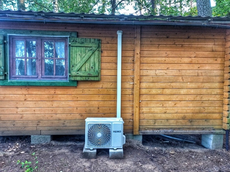 Biała jednostka zewnętrzna klimatyzatora marki Heiko, zamontowana na betonowych bloczkach przy drewnianej ścianie domku letniskowego z zielonymi okiennicami i widoczną rurą odprowadzającą skropliny.