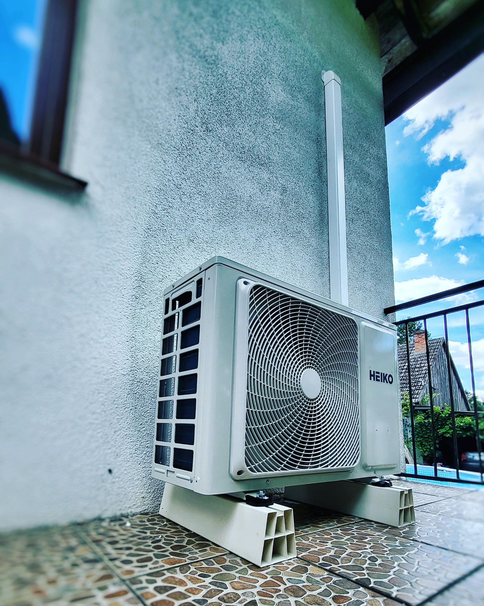 Zewnętrzna jednostka klimatyzacji marki HEIKO zamontowana na balkonie, widoczna instalacja odprowadzająca skropliny, w tle błękitne niebo i fragment budynku.