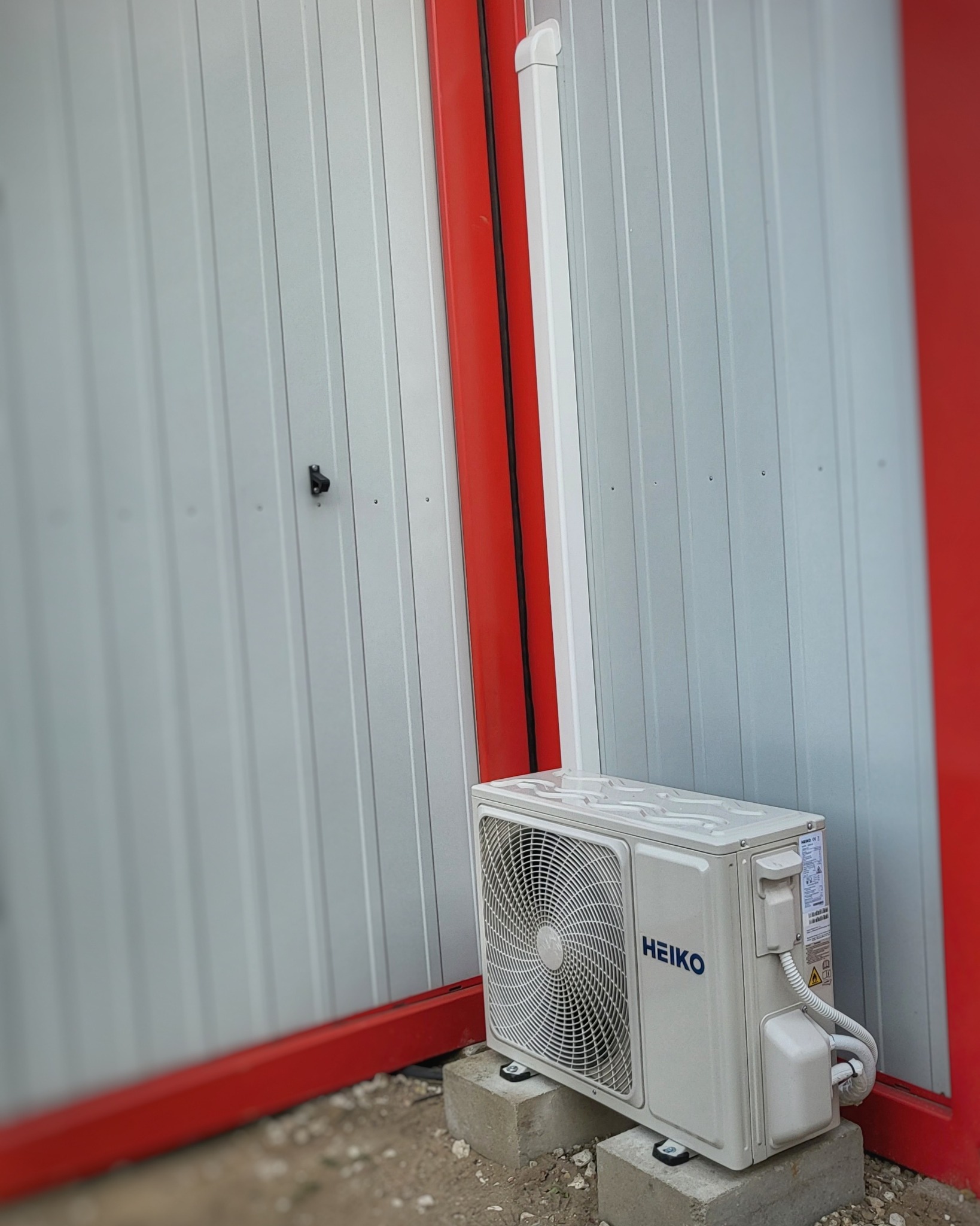 Zewnętrzna jednostka klimatyzacji marki Heiko, ustawiona na betonowych bloczkach przy szarej ścianie z czerwonymi elementami konstrukcyjnymi i białą rurą spustową.