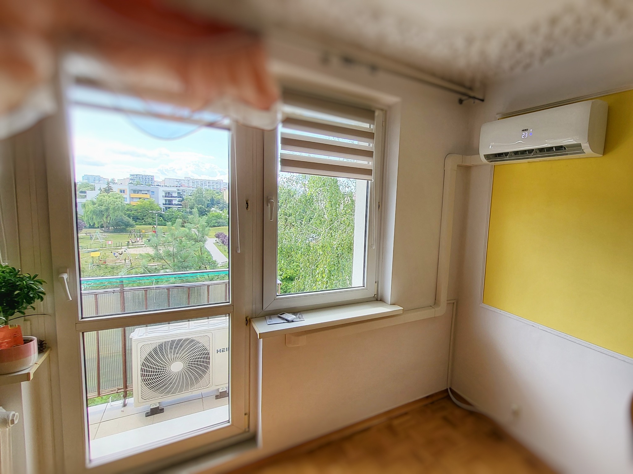 Wnętrze pokoju z klimatyzatorem ściennym HEIKO, widok przez okno na balkon z jednostką zewnętrzną klimatyzacji i panoramę osiedla z placem zabaw.