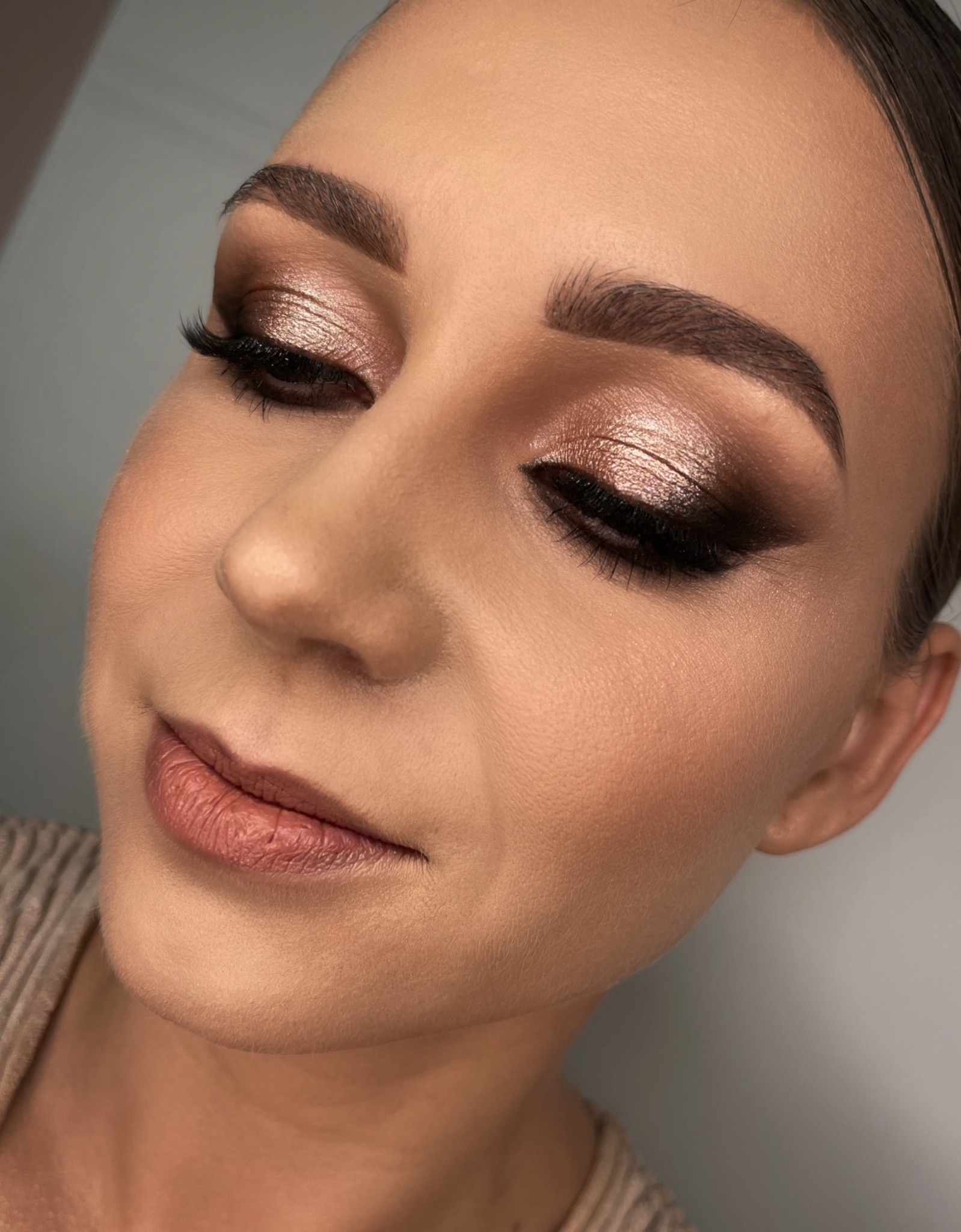 Portret kobiety z profesjonalnym, błyszczącym makijażem oczu w odcieniach brązu i beżu, podkreślonym eyelinerem i sztucznymi rzęsami, oraz delikatnym makijażem ust w kolorze nude.
