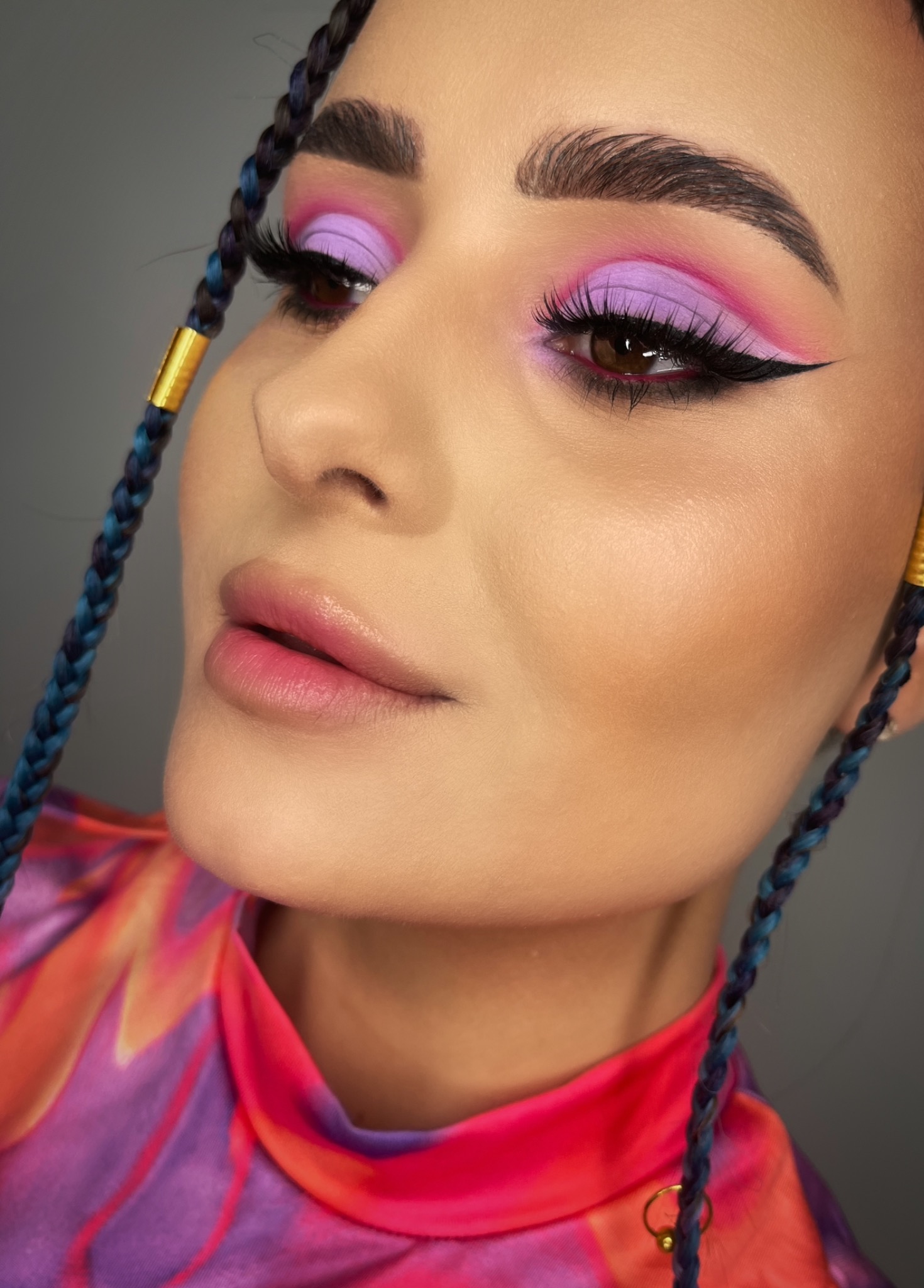 Portret kobiety z intensywnym makijażem oczu w odcieniach fioletu i różu, czarnym eyelinerem i sztucznymi rzęsami, z niebiesko-fioletowymi warkoczykami ozdobionymi złotymi pierścieniami.