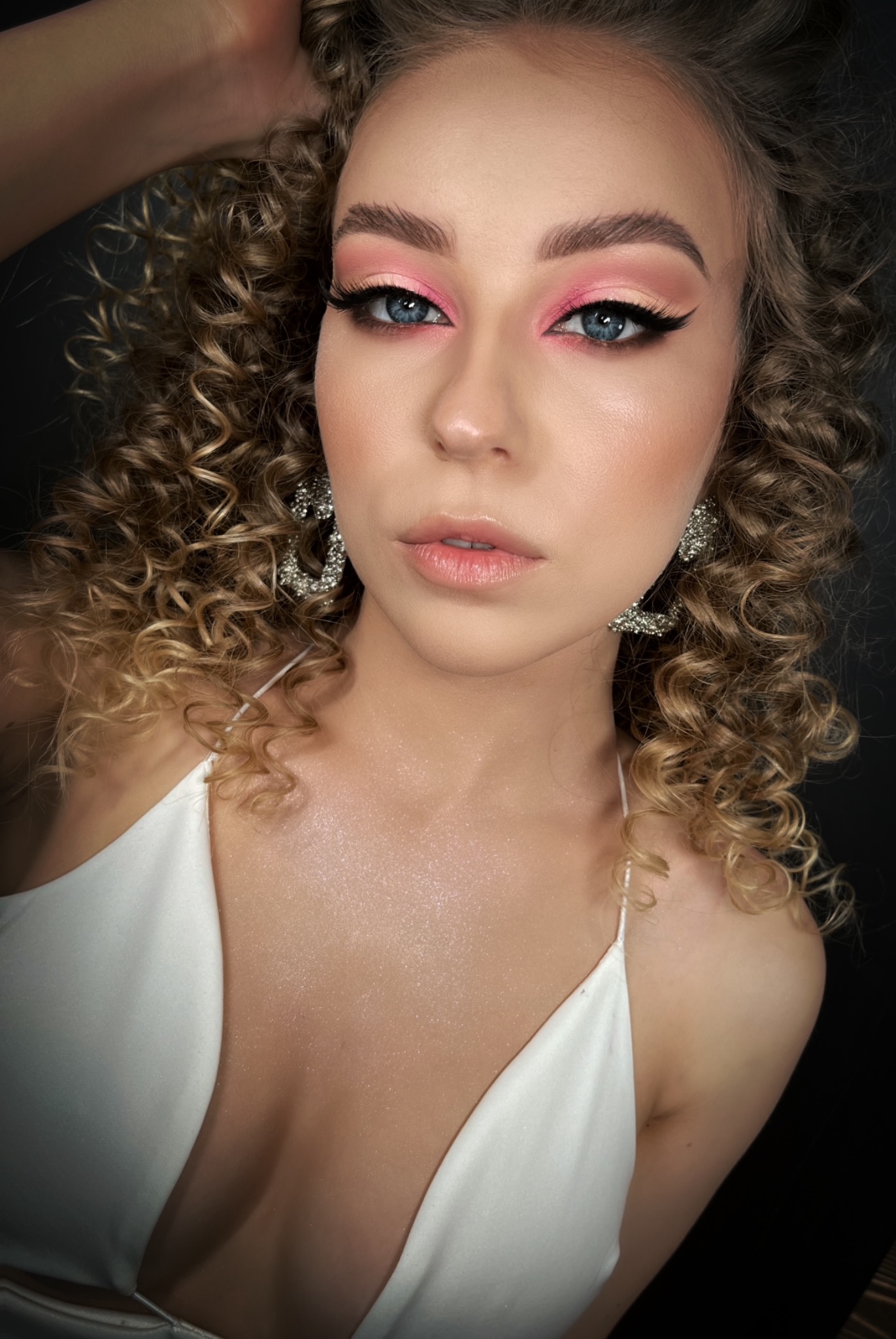 Portret młodej kobiety z profesjonalnym, różowym makijażem oczu, podkreślonym eyelinerem, kręconymi blond włosami i błyszczącymi kolczykami na tle czerni.