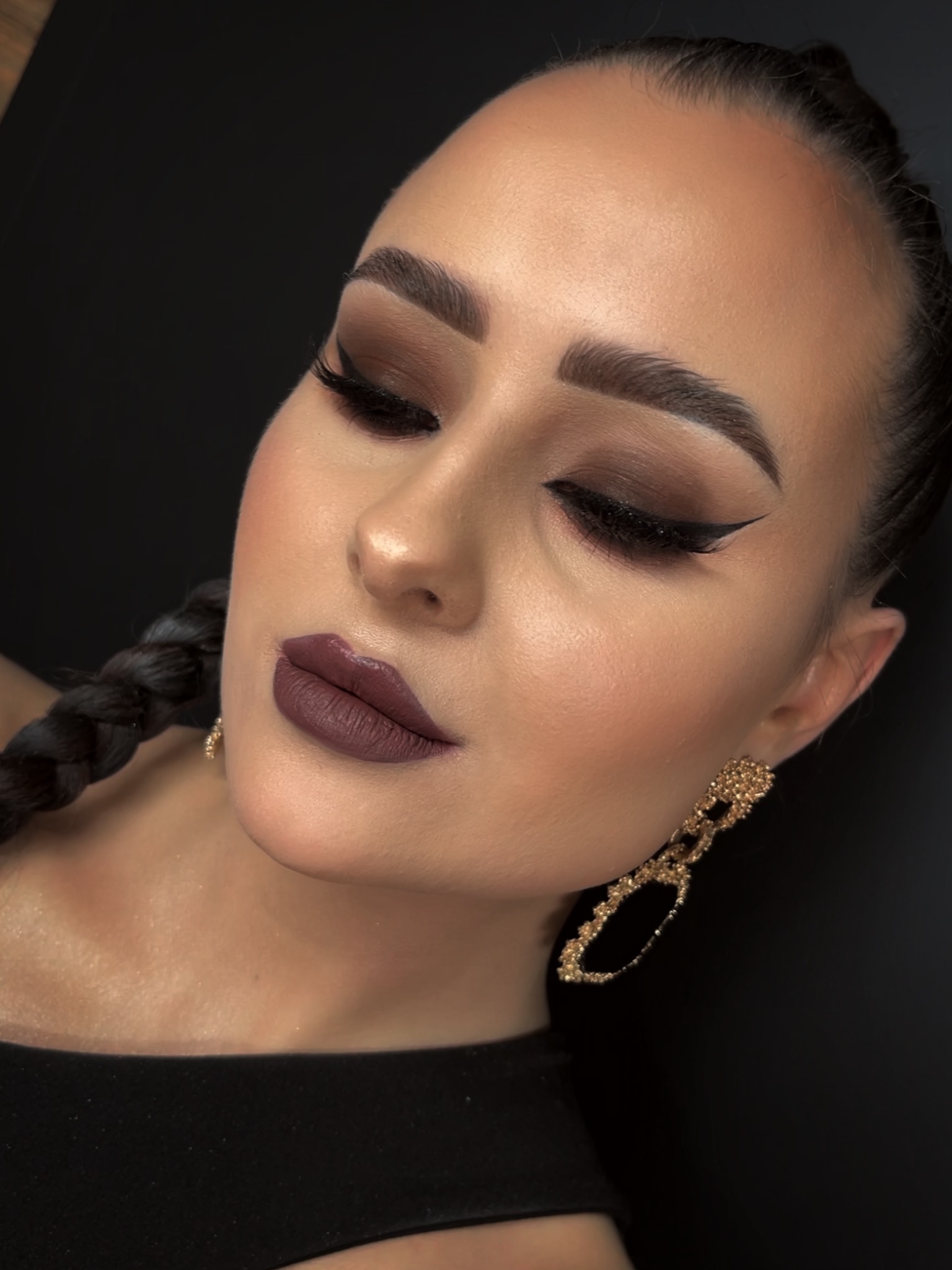 Portret kobiety z profesjonalnym, wieczorowym make-up'em: brązowe cienie, czarny eyeliner, ciemna pomadka, podkreślone brwi i złote kolczyki, włosy splecione w warkocz.