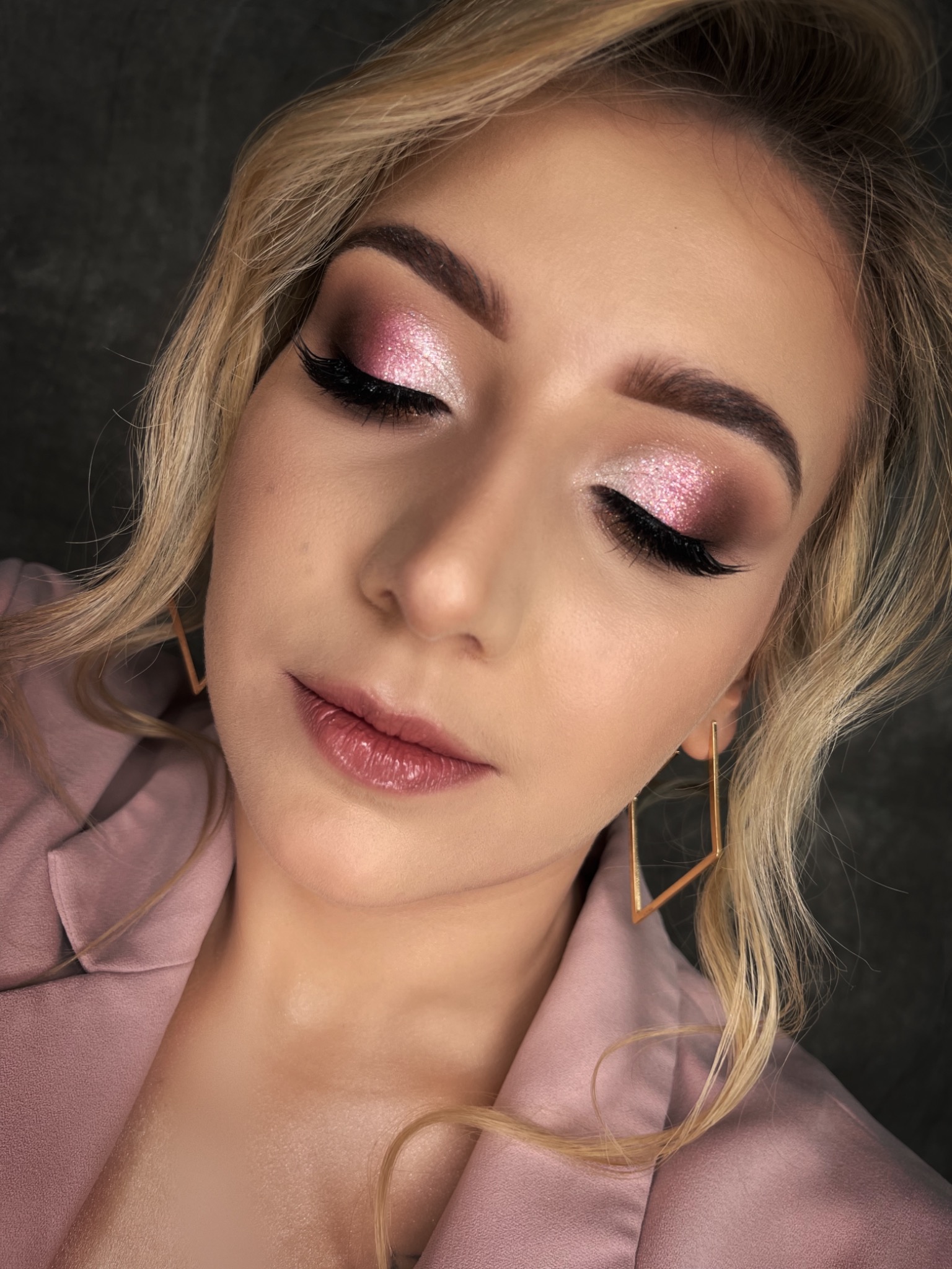 Portret kobiety z zamkniętymi oczami, prezentującej błyszczący, różowo-brązowy cień do powiek, długie rzęsy, błyszczące usta i geometryczne, złote kolczyki na tle ciemnej ściany i różowej marynarki.