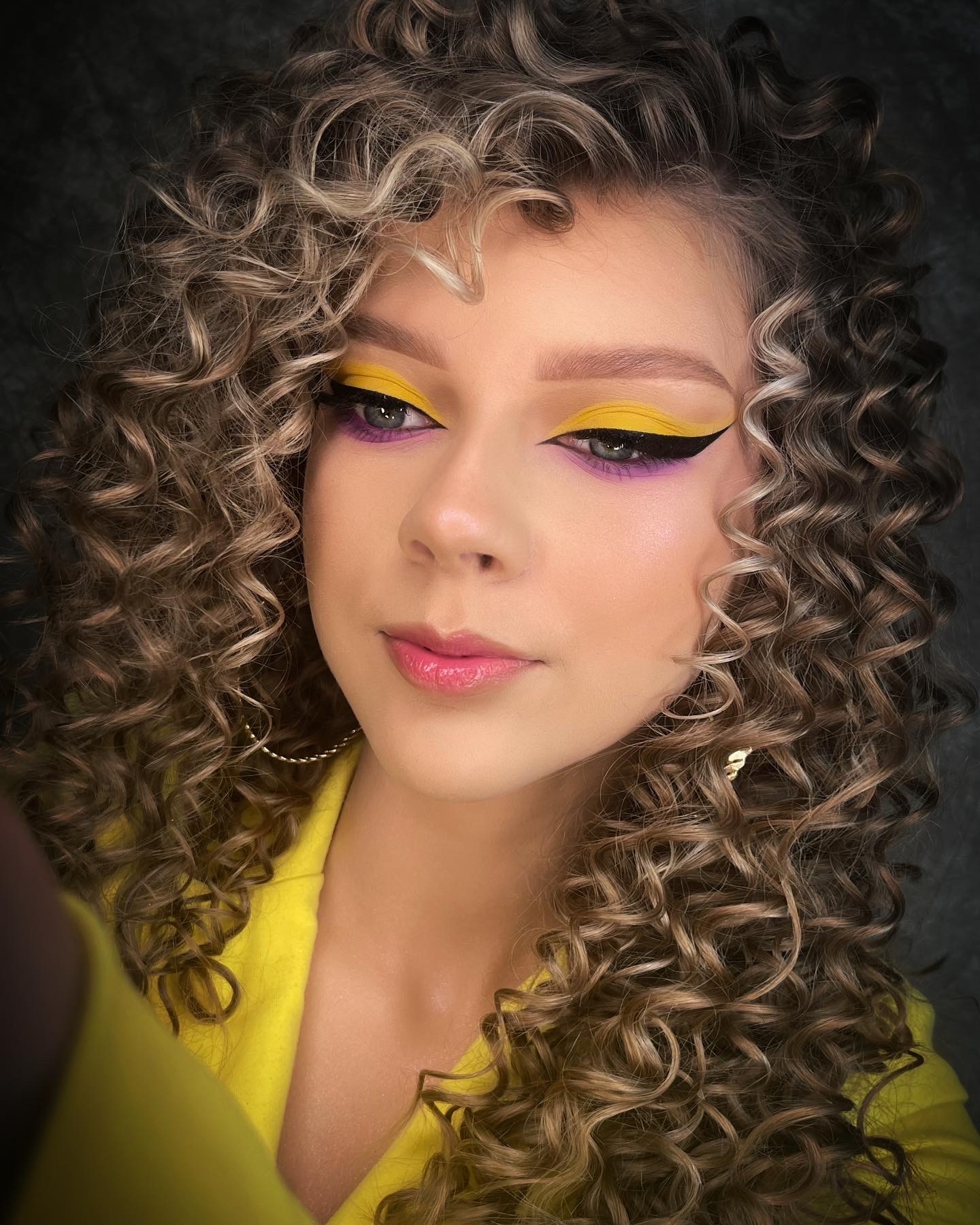 Portret kobiety z burzą loków, prezentującej kreatywny makijaż oczu: żółty cień na powiekach z czarną kreską i fioletowym akcentem pod dolną powieką, subtelny błyszczyk na ustach, złote kolczyki koła.