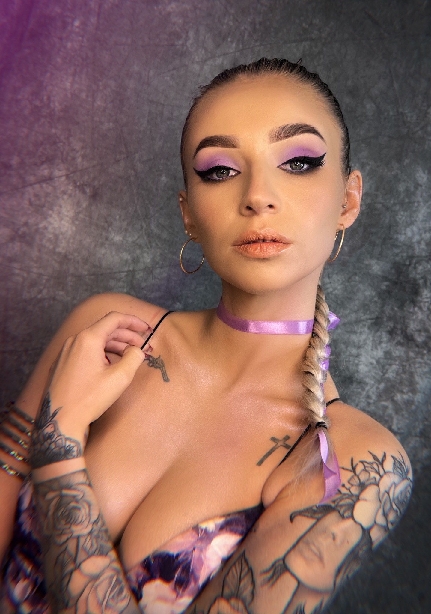 Portret kobiety z wyrazistym fioletowym cieniem do powiek, czarnym eyelinerem, i naturalnymi ustami, włosy zaplecione w warkocz z fioletową wstążką, widoczne tatuaże na ramionach i dekolcie, w tle...