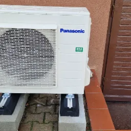 Zewnętrzna jednostka klimatyzacyjna Panasonic R32, zamontowana na betonowych bloczkach i gumowych podkładkach, stoi na kostce brukowej obok brązowych drzwi wejściowych.