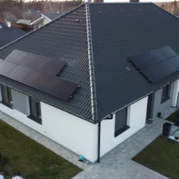 Widok z góry na nowoczesny dom jednorodzinny z ciemną dachówką i zamontowanymi panelami słonecznymi na dachu, otoczony zadbanym trawnikiem i brukowanym chodnikiem.