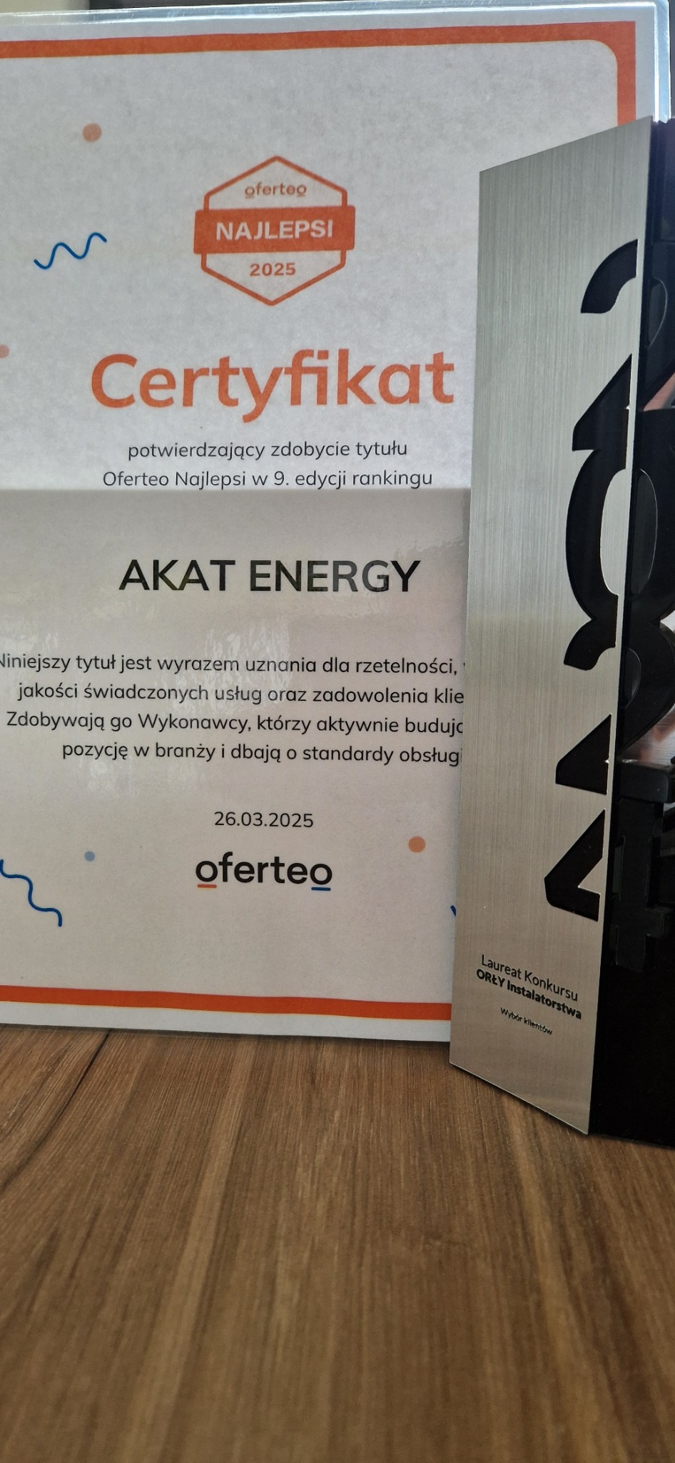 Certyfikat Oferteo Najlepsi 2025 dla AKAT ENERGY oraz statuetka 'Orły Instalatorstwa'. Uznanie za rzetelność i jakość usług. Dekoracja na drewnianym podłożu.