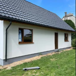 Świeżo pomalowana, biała elewacja domu jednorodzinnego z brązowymi oknami i drzwiami, czarny dach, rynna i szara podmurówka, widoczny fragment trawnika z rurą PCV.