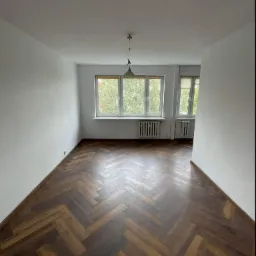 Pusty pok&oacute;j z parkietem w jodełkę, świeżo pomalowanymi białymi ścianami i dwoma oknami z widokiem na zieleń.