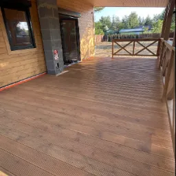 Drewniany taras z ryflowaną deską, widok na ogród przez balustradę, surowy komin z bloczków betonowych przy ścianie z drewnianą boazerią.