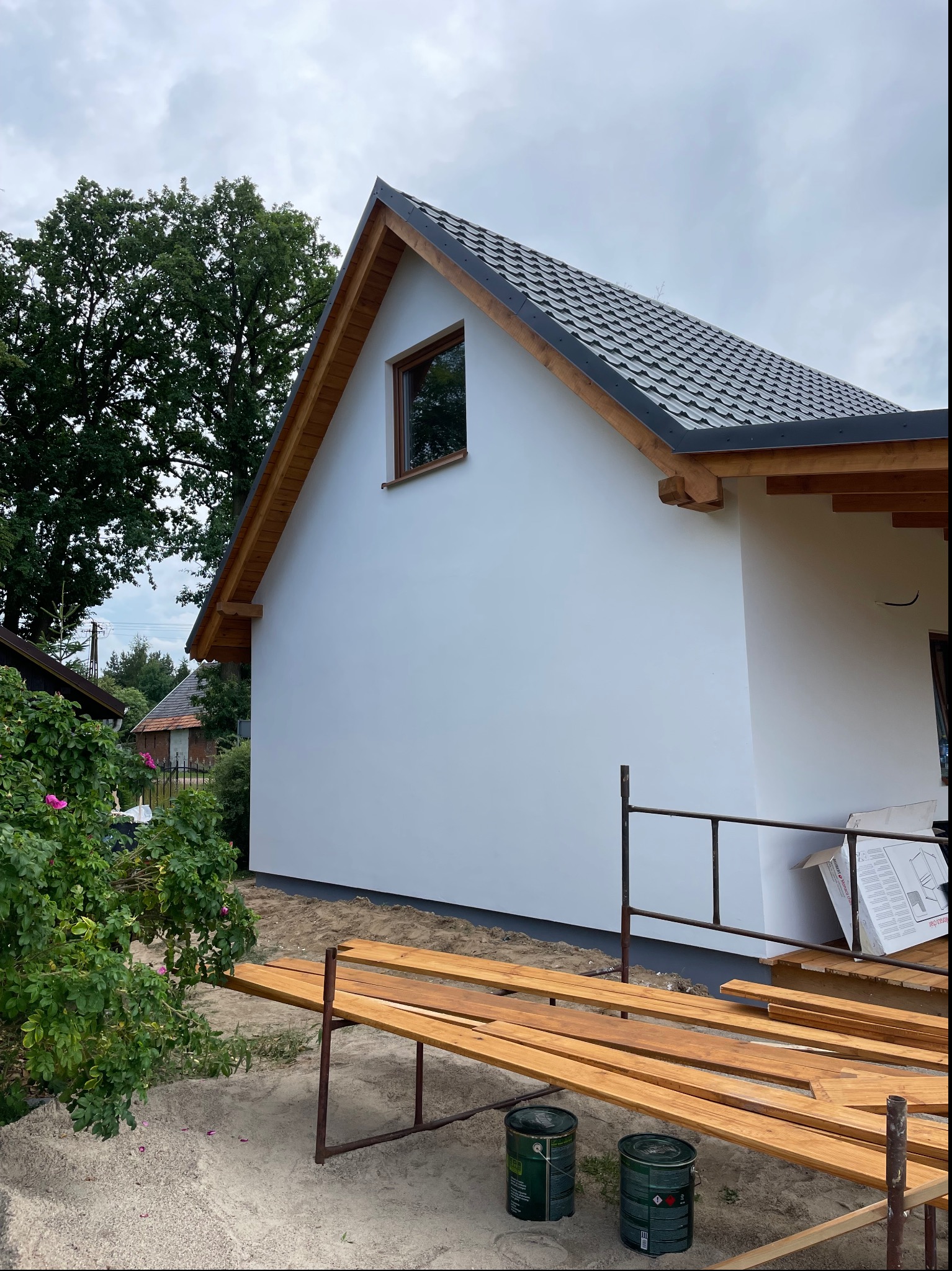 Świeżo pomalowana, biała elewacja domu jednorodzinnego z szarą dachówką, widoczne rusztowanie, drewniane deski i puszki z farbą, drzewo w tle.