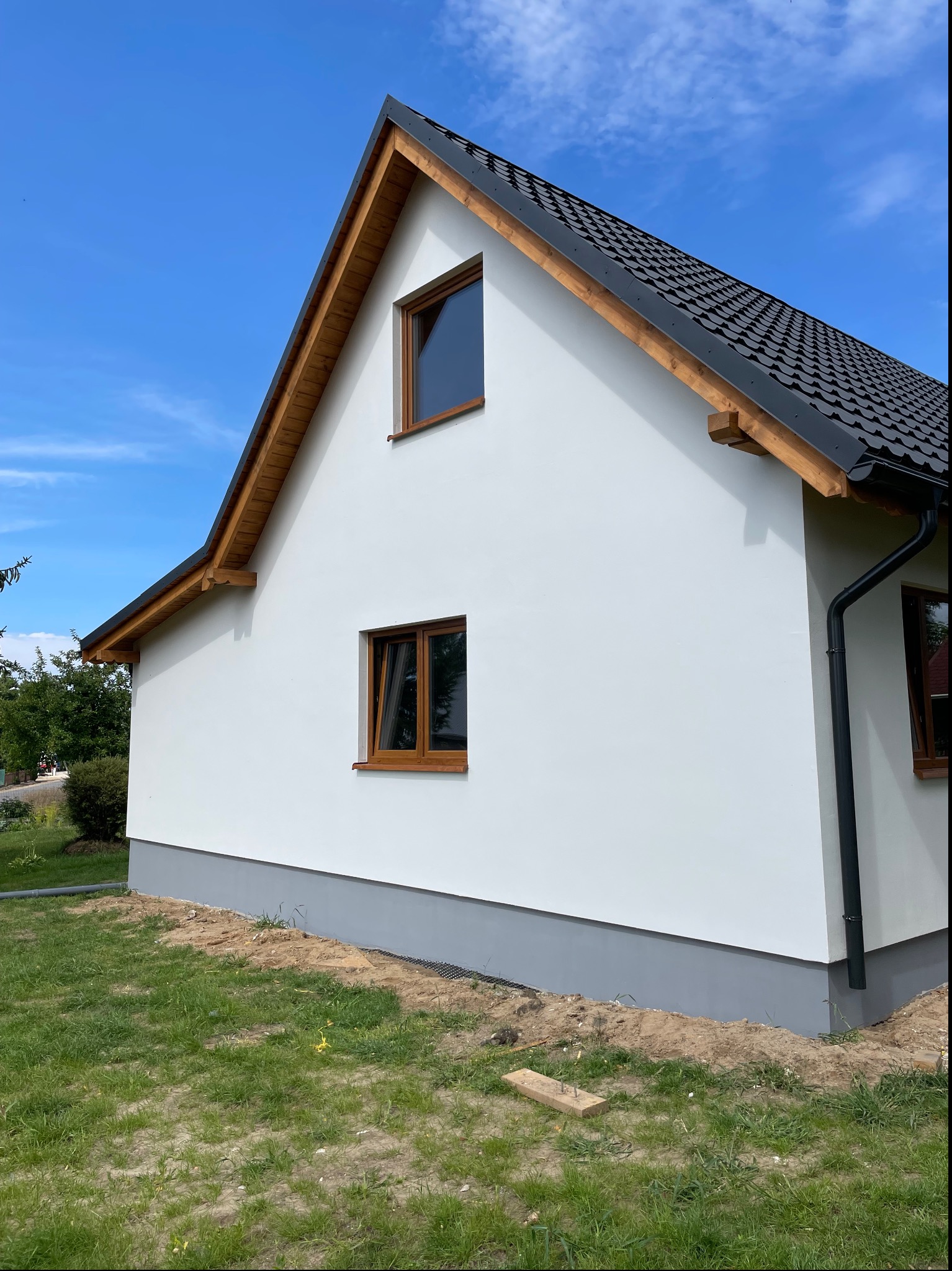 Świeżo otynkowany dom jednorodzinny z białymi ścianami i szarą podmurówką, widoczne drewniane elementy wykończenia dachu i brązowe ramy okienne, teren wokół domu częściowo zagospodarowany trawą...