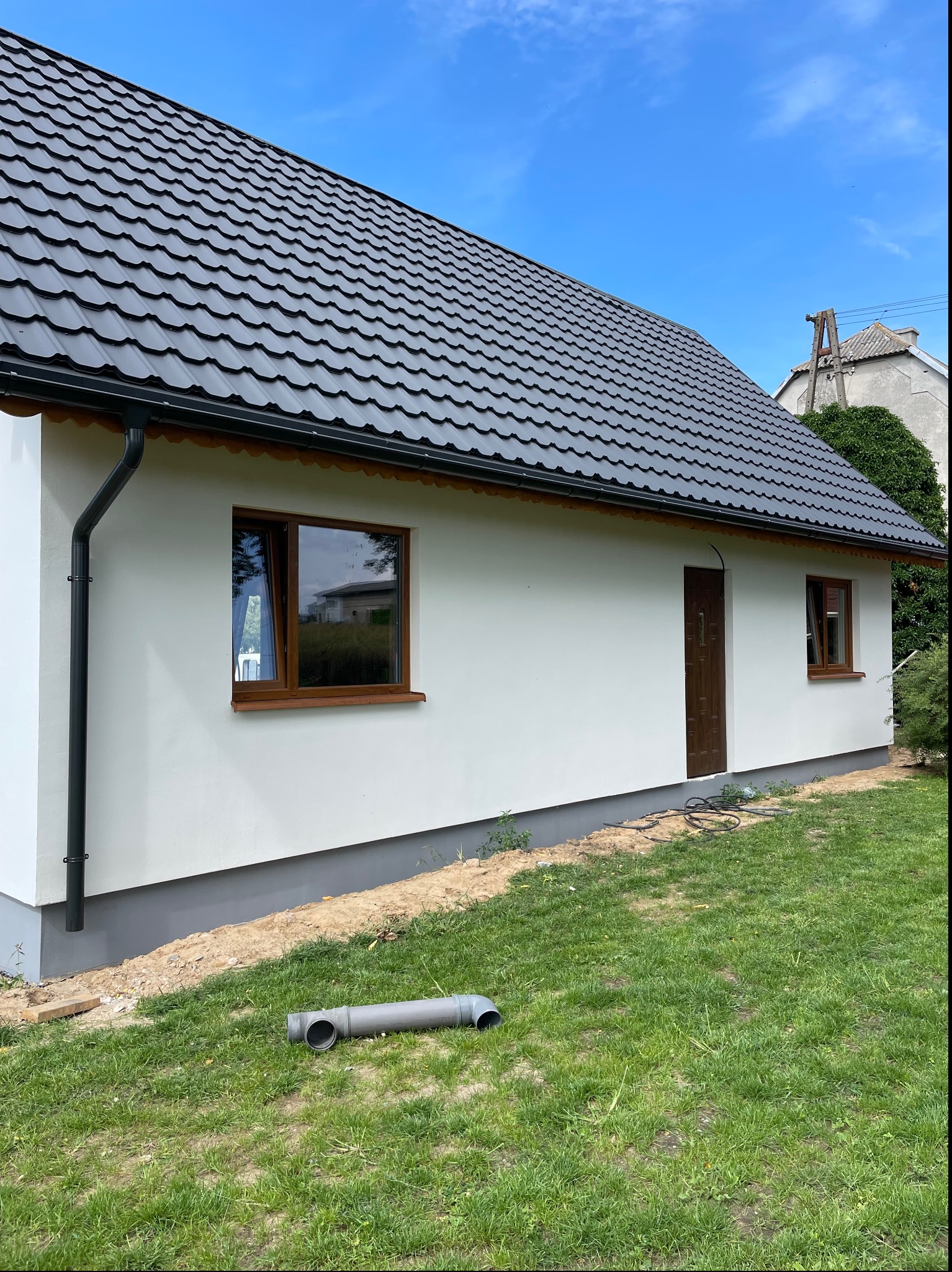 Świeżo pomalowana, biała elewacja domu jednorodzinnego z brązowymi oknami i drzwiami, czarny dach, rynna i szara podmurówka, widoczny fragment trawnika z rurą PCV.