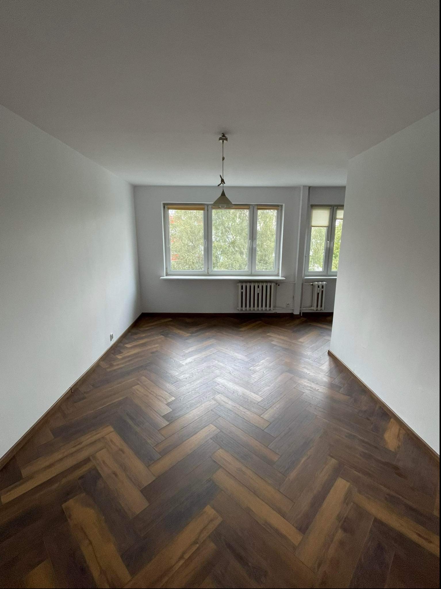 Pusty pokój z parkietem w jodełkę, świeżo pomalowanymi białymi ścianami i dwoma oknami z widokiem na zieleń.
