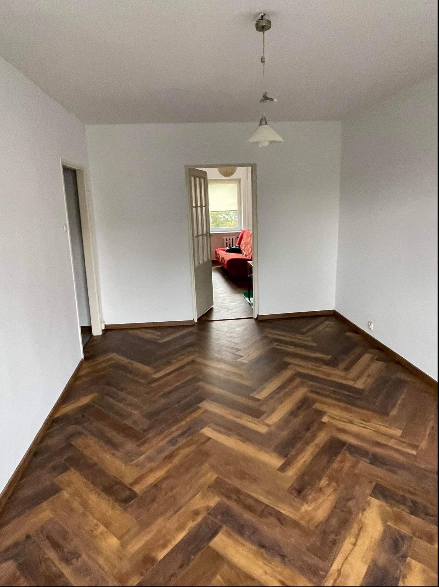 Wnętrze pokoju w Jabłonowie Pomorskim z ułożoną podłogą z paneli w jodełkę, świeżo pomalowane ściany na biało, widok na pokój z czerwonym narożnikiem przez otwarte drzwi.