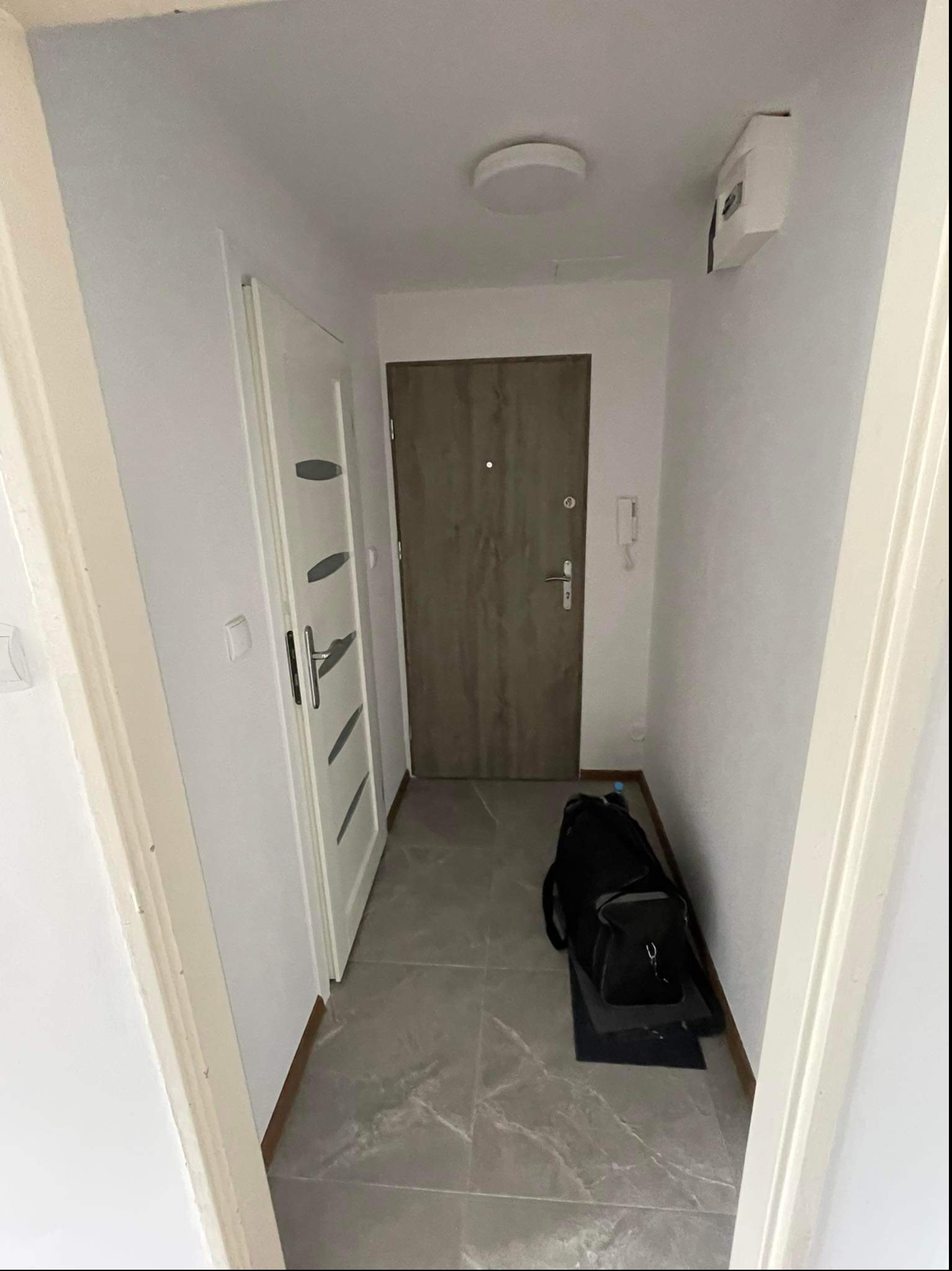 Wnętrze przedpokoju z szarą podłogą, białą ścianą i drzwiami wejściowymi, widoczna torba podróżna.