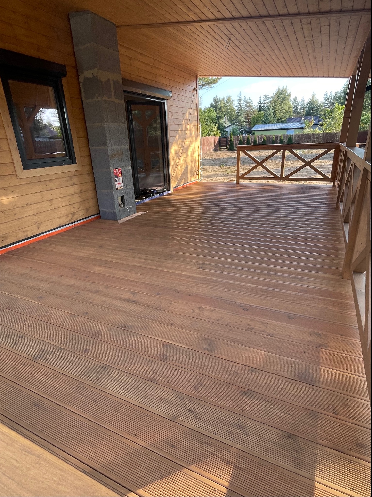 Drewniany taras z ryflowaną deską, widok na ogród przez balustradę, surowy komin z bloczków betonowych przy ścianie z drewnianą boazerią.