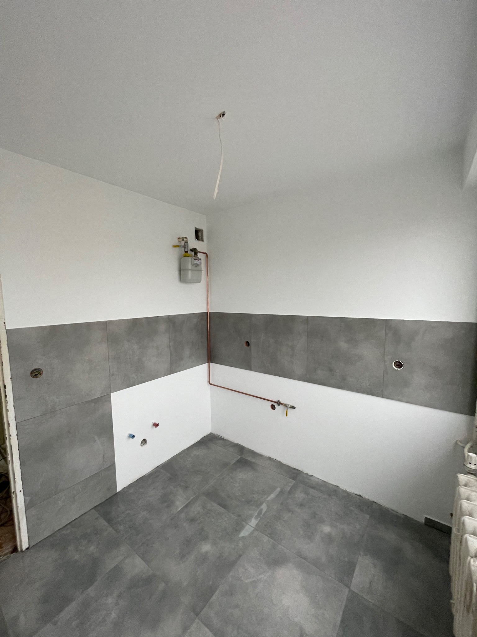 Łazienka w trakcie wykańczania: ułożone szare płytki podłogowe i częściowo na ścianach, widoczne instalacje wodne i gazowe, przygotowane otwory pod armaturę, białe ściany powyżej płytek.