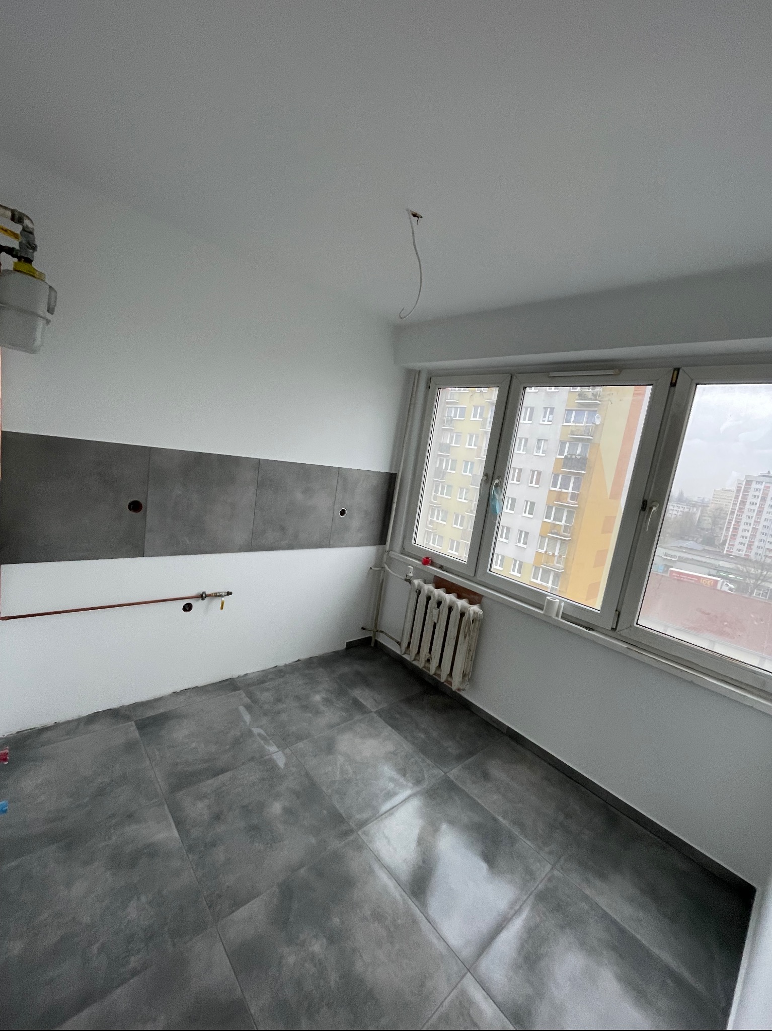 Wnętrze pokoju po remoncie, z szarymi płytkami na podłodze i częściowo na ścianie, widoczne rury i grzejnik przy oknie z widokiem na blok.