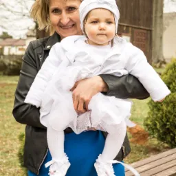 Uśmiechnięta kobieta trzyma na rękach dziewczynkę w białym ubranku i czapeczce z kokardką, w tle drewniana altana i ławka.