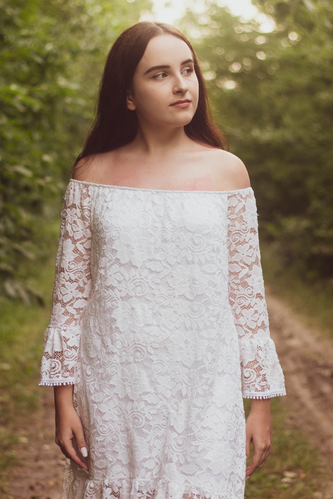 Młoda kobieta w białej koronkowej sukience z odkrytymi ramionami, stojąca na leśnej ścieżce, patrzy w dal.
