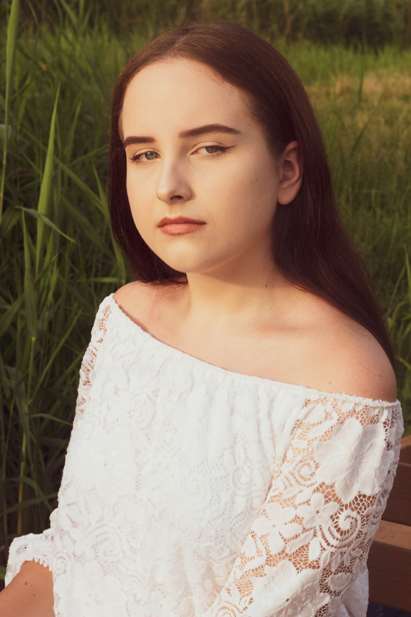 Portret młodej kobiety w białej koronkowej bluzce z odkrytymi ramionami, siedzącej na drewnianej ławce na tle zielonej trawy, z delikatnym makijażem i poważnym wyrazem twarzy.