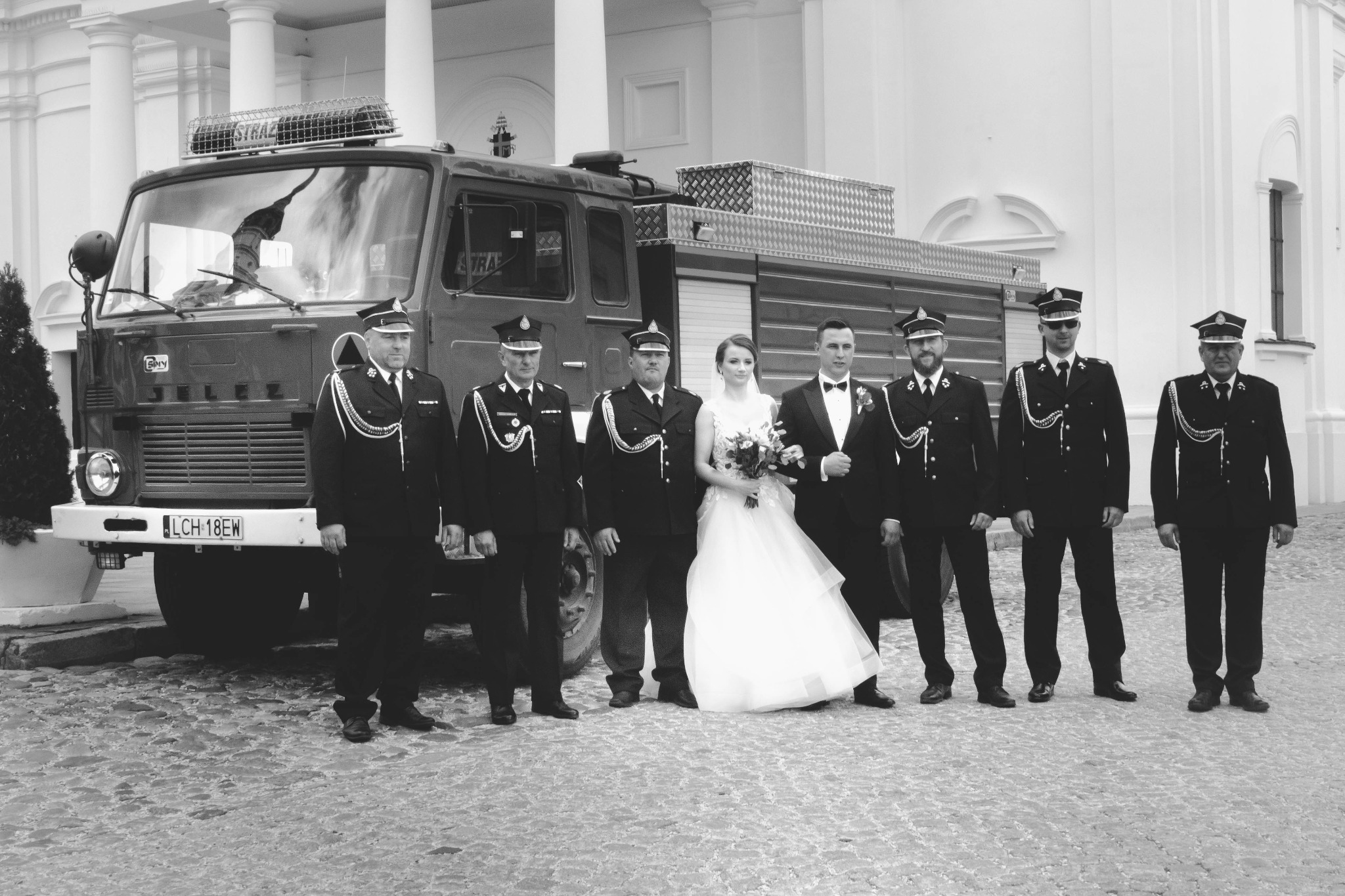 Czarno-białe zdjęcie ślubne: para młoda i strażacy w mundurach pozują przed wozem strażackim w tle kolumnady.