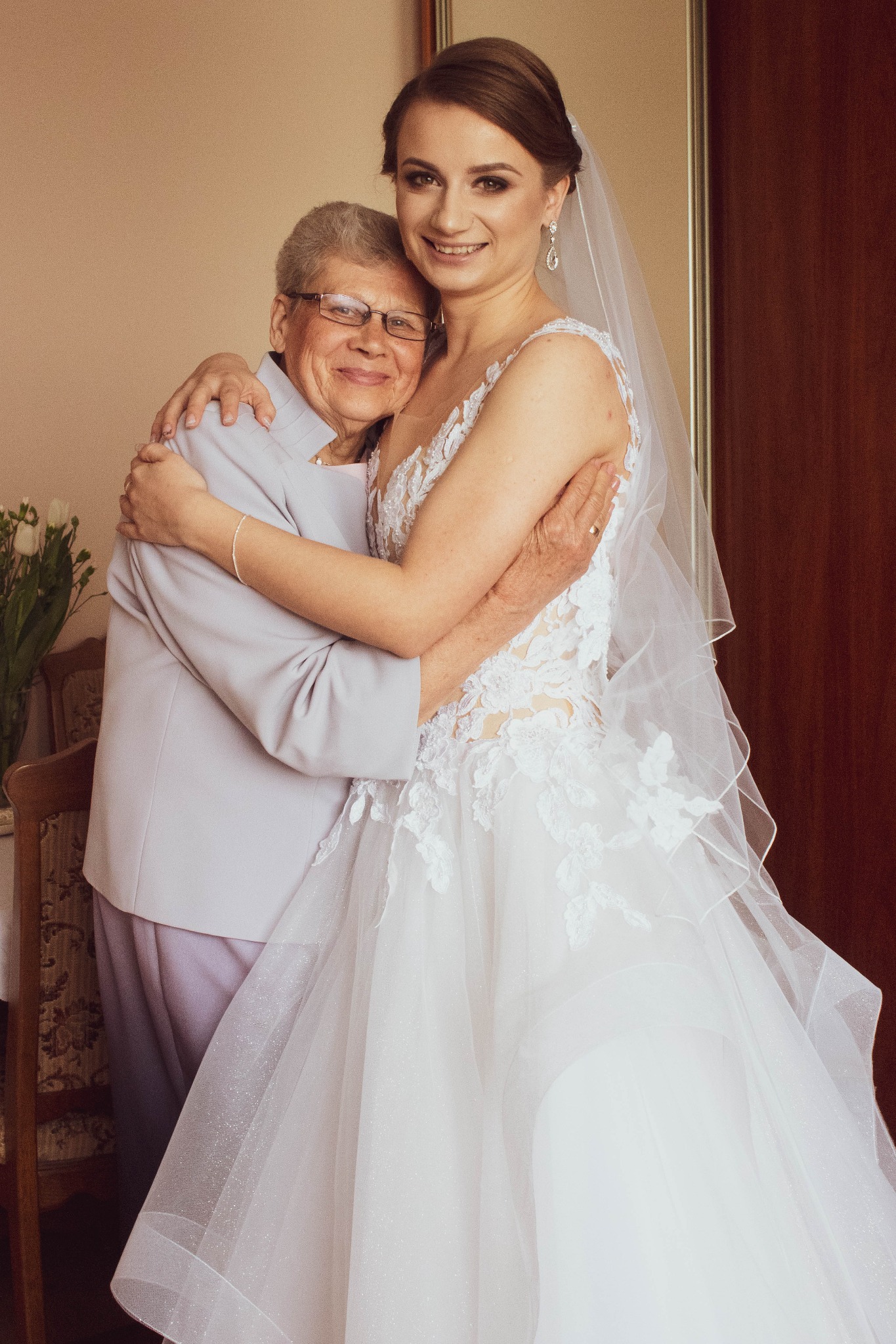Uśmiechnięta panna młoda w białej sukni ślubnej z koronkowymi aplikacjami, obejmująca starszą kobietę w pastelowym garniturze, podczas przygotowań do ceremonii.