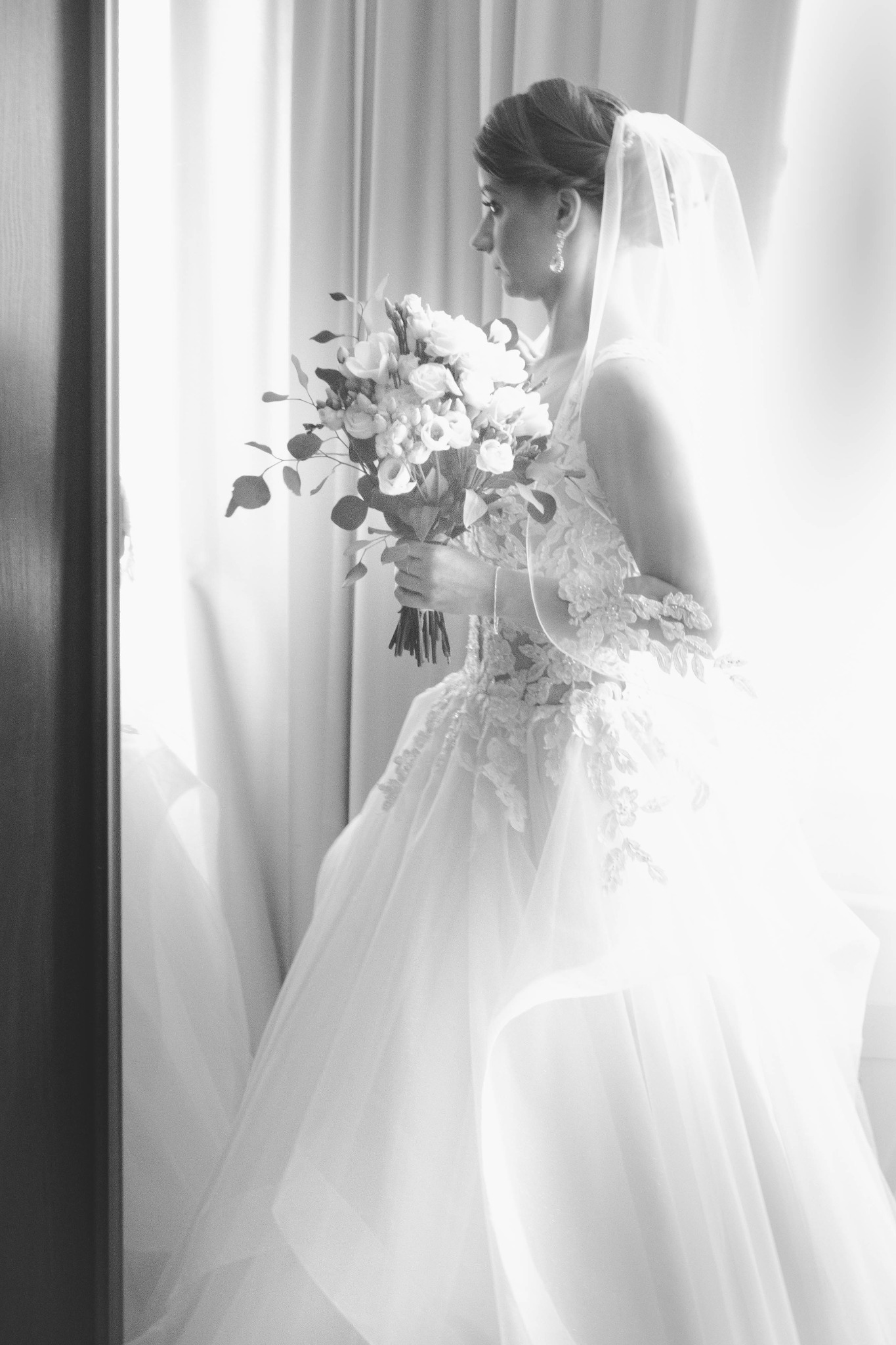 Czarno-białe zdjęcie panny młodej w sukni ślubnej z bukietem kwiatów, stojącej przy oknie z zasłonami, widok z profilu.
