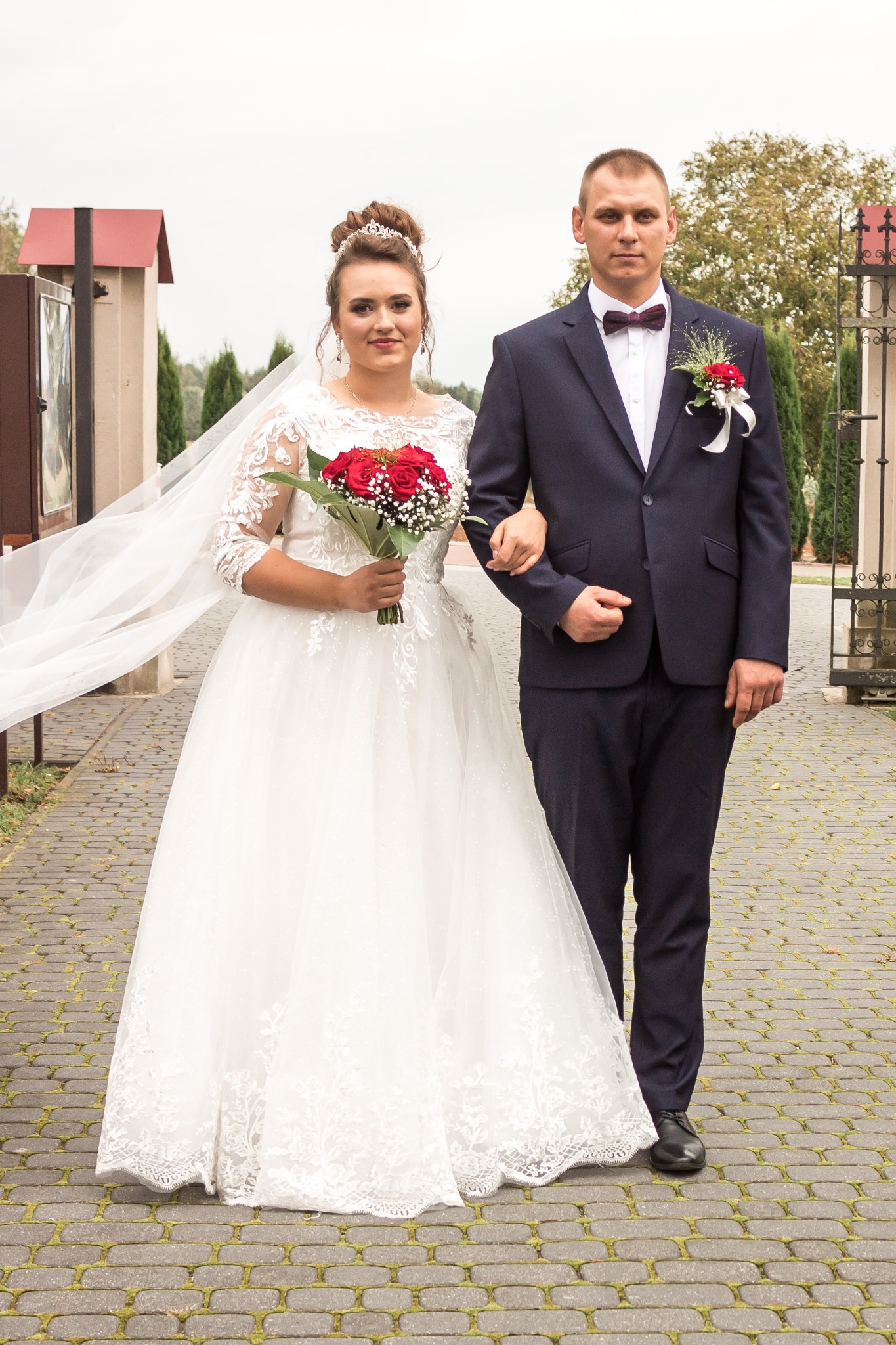 Para młoda: panna młoda w białej sukni z koronkowymi rękawami trzyma bukiet czerwonych róż, a pan młody w granatowym garniturze z bordową muchą, stoją na brukowanej ścieżce.